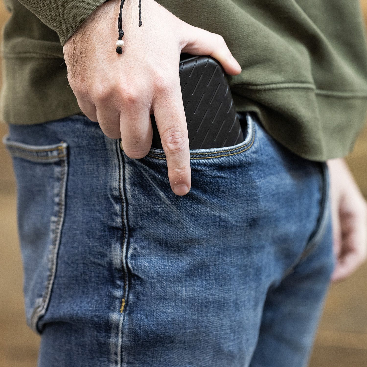 Harrows Slimcase Darttasche Eine Person steckt ein schwarzes Etui in die Hosentasche. Sie trägt eine grüne Jacke und eine blaue Jeans.
