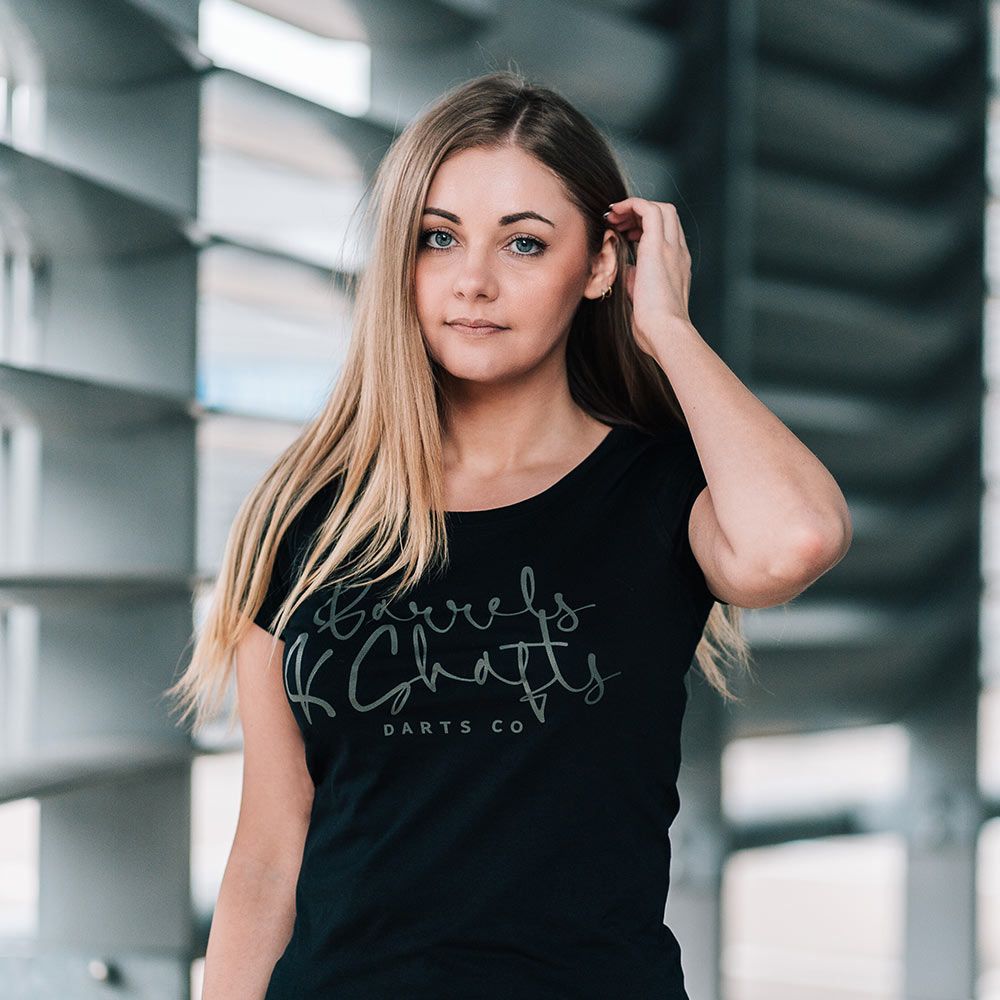 Barrels and Shafts Damen T-Shirt - Schwarz Eine Frau trägt ein schwarzes T-Shirt mit der Aufschrift „Barrels and Shafts Darts Co“. Sie steht vor einem modernen, lichtdurchfluteten Hintergrund.