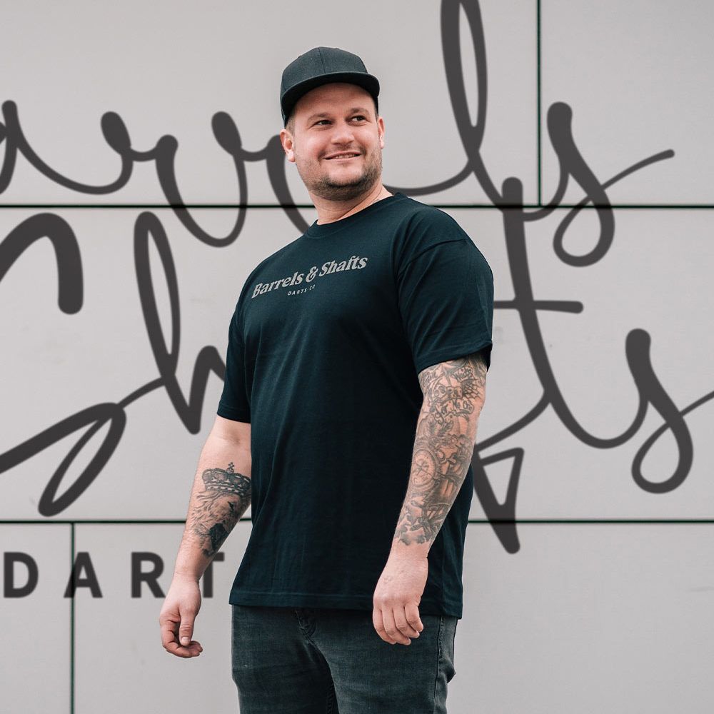 Barrels and Shafts T-Shirt - Schwarz Ein Mann trägt ein schwarzes T-Shirt mit der Aufschrift „Barrels & Shafts“. Er steht vor einer Wand mit passender Schrift im Hintergrund.