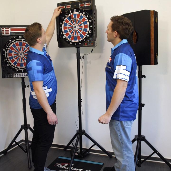 Zwei Männer in blauen Shirts stehen vor elektronischen Dartscheiben. Einer der Männer nimmt einen Dartpfeil aus der Dartscheibe.